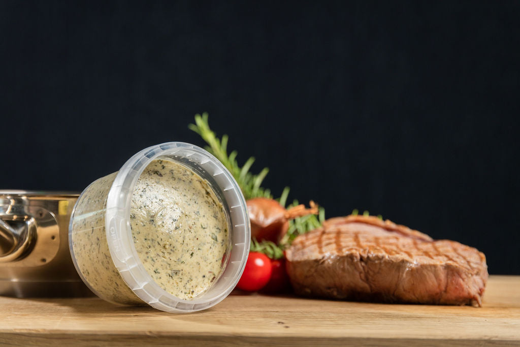 Cafe de Paris Kräuterbutter von Degusta Manufaktur im Becher, daneben Rosmarin, eine Kirschtomate und ein grilliertes Rindssteak